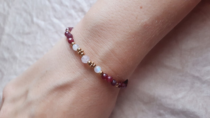 May include: A beaded bracelet featuring small, faceted, deep red gemstones and white, round beads. Gold-colored beads separate the gemstones. The bracelet is worn on a wrist.