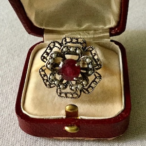 Peut inclure: Une bague en or avec une pierre rouge et un motif floral orné de perles blanches. La bague est présentée dans un écrin à bijoux en velours rouge.