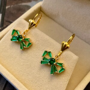 Peut inclure: Une paire de boucles d'oreilles en or avec des accents de nœud en cristal vert. Les boucles d'oreilles sont présentées dans une boîte en velours beige.