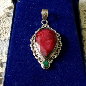Peut inclure: Un pendentif en argent avec une grande pierre précieuse rouge et une plus petite pierre précieuse verte. Le pendentif a un design délicat en filigrane.