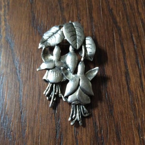 Peut inclure: Broche argentée représentant un groupe de trois fleurs de fuchsia stylisées avec des feuilles détaillées. La broche est un bijou délicat et complexe.