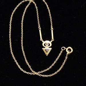 Peut inclure: Un collier en chaîne dorée avec un pendentif géométrique en forme de triangle noir surmonté d'une pierre rouge.