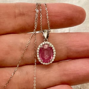 May include: A silver necklace with a pendant featuring a pink oval gemstone, encircled by small, clear stones. The pendant is suspended from a fine silver chain, presenting a classic and elegant design. The gemstone has a vibrant colour.