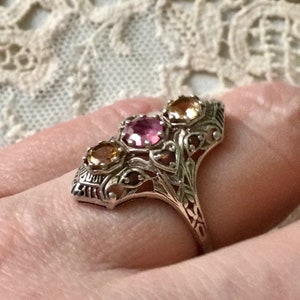 Peut inclure: Une bague en argent avec un motif de filigrane complexe présente trois pierres précieuses : une pierre centrale rose et deux pierres plus petites de couleur ambre de chaque côté.