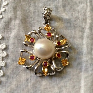 May include: A silver and gold pendant featuring a large white pearl surrounded by smaller red and gold stones. The pendant is shaped like a flower with intricate details.