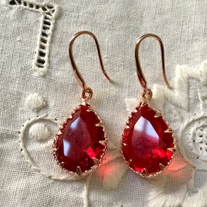 Peut inclure: Deux boucles d'oreilles en forme de goutte d'eau avec des pierres précieuses rouge vif. Les boucles d'oreilles ont un sertissage et des crochets d'oreille en métal de couleur or rose. Les pierres précieuses sont facettées et bordées d'un bord festonné délicat.