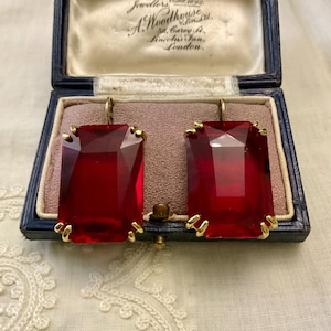 Peut inclure: Paire de boucles d'oreilles rectangulaires en pierres précieuses rouges, serties de métal doré. Elles sont présentées dans un écrin bleu ouvert, avec un intérieur rose. Le coffret porte une inscription.