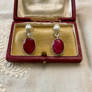 Peut inclure: Boucles d'oreilles en argent avec des pierres précieuses ovales rouge foncé et de petites perles blanches. Les boucles d'oreilles sont présentées dans un écrin à bijoux rouge vintage avec un intérieur doré.