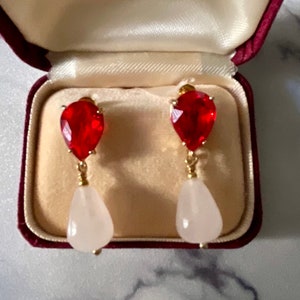 Peut inclure: Une paire de boucles d'oreilles en forme de goutte d'eau rouge et blanche avec des accents dorés. Les boucles d'oreilles sont dans une boîte en velours rouge.