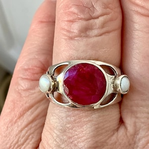 Peut inclure: Une bague en argent ornée d'une grande pierre précieuse ronde à facettes rouge foncé au centre, flanquée de deux petites pierres cabochons rondes et irisées blanches de chaque côté. L'anneau présente un design ajouré autour des pierres.