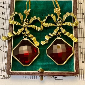 Peut inclure: Une paire de boucles d'oreilles en or avec une pierre précieuse rouge facetée. Les boucles d'oreilles présentent un motif de nœud en or.