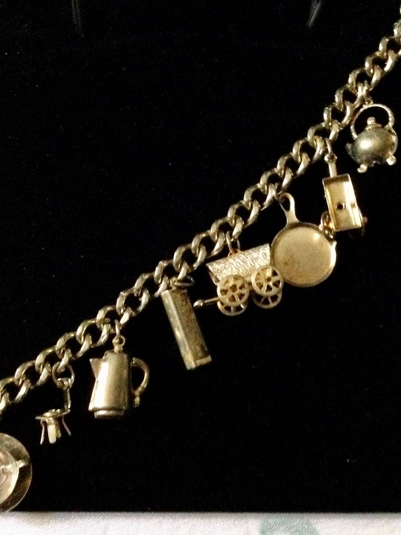 Pode incluir: Um bracelete de ouro com v&aacute;rios pingentes, incluindo um bule, uma carro&ccedil;a, uma frigideira, uma caixa de f&oacute;sforos e um jarro.