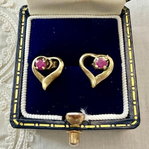 Peut inclure: Boucles d'oreilles en forme de cœur en or, chacune ornée d'un petit rubis rond. Les boucles d'oreilles sont présentées dans un écrin à bijoux bleu vintage avec une fermeture et une bordure dorées.