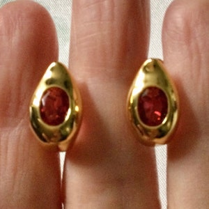 Peut inclure: Une paire de boucles d'oreilles en forme de goutte dorée, chacune présentant une finition métallique lisse et polie avec une pierre précieuse rouge foncé de taille ovale sertie au centre, offrant un aspect réfléchissant et audacieux.