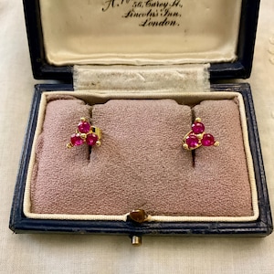 May include: A pair of gold stud earrings, each featuring three round, vibrant pinkish-red gemstones arranged in a triangular cluster. The earrings are presented inside a dark blue, hinged jewellery box with a cream-coloured interior lining and the printed text: Jewellers Estd. 1692. A. Woodhouse & Son, Ltd. 56, Carey St., Lincoln's Inn, London.