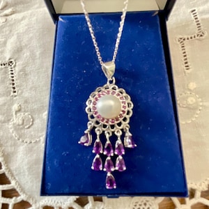 May include: A silver necklace with a pendant featuring a large, round, white pearl surrounded by small pink stones. The pendant has a decorative silver filigree design and dangling purple teardrop-shaped stones. The necklace is displayed in a blue jewellery box.