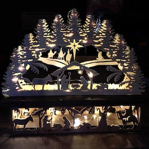May include: A white wooden Christmas village silhouette with a nativity scene and a star on top. The village is illuminated with warm white lights.