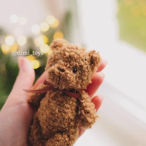 May include: A small, brown, fuzzy teddy bear with a textured, curly surface, dark bead eyes, and a stitched nose. The bear features a thin, dark red ribbon tied around its neck. The text @dimi_toys is visible on the left side of the image.