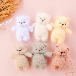 May include: Six small, fuzzy, hand-knitted teddy bears arranged in two rows. The bears are colored in light purple, white, pale yellow, beige, light gray, and soft pink. Each bear features embroidered black eyes and a nose, with the text @dimi_toys visible.
