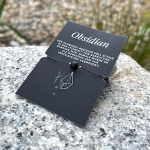 Puede incluir: Un collar de cuentas de obsidiana negra con una etiqueta negra. La etiqueta dice "Obsidian" e incluye texto en alemán. El collar se muestra sobre una superficie de piedra gris.
