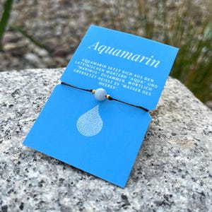 Könnte beinhalten: Eine blaue Karte mit dem Wort "Aquamarin" und deutschem Text liegt auf einem Stein. Ein schwarzes Armband mit einer hellblauen Perle und goldenen Akzenten liegt vor der Karte. Die Karte hat eine weiße Wassertropfen-Grafik.