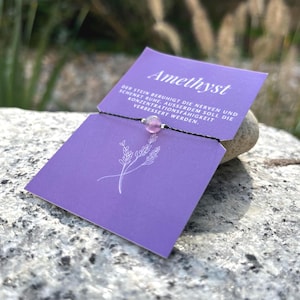 Könnte beinhalten: Nahaufnahme eines Armbands mit einer Amethystperle an einem schwarzen Band, präsentiert auf einer lila Karte mit dem Wort "Amethyst" und deutschem Text. Die Karte liegt auf einem grauen Stein, mit verschwommenem natürlichem Hintergrund.