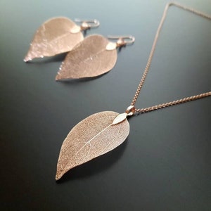 May include: A delicate rose gold necklace and earrings set featuring leaf-shaped pendants. The leaves are made of thin, intricate metalwork and have a delicate, airy appearance.