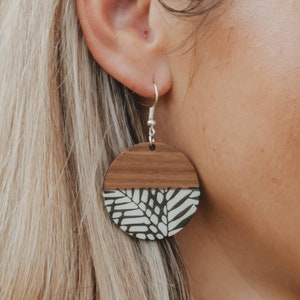 Peut inclure: Une paire de boucles d'oreilles rondes en bois avec un motif de feuilles noires et blanches. Les boucles d'oreilles sont suspendues à des crochets argentés.