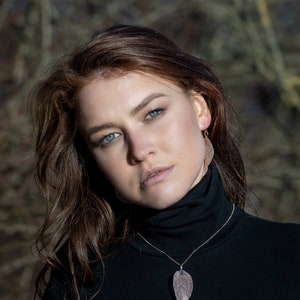 May include: A woman with long brown hair wearing a black turtleneck sweater with a gold necklace featuring a leaf pendant. The leaf pendant is made of a sparkly material.