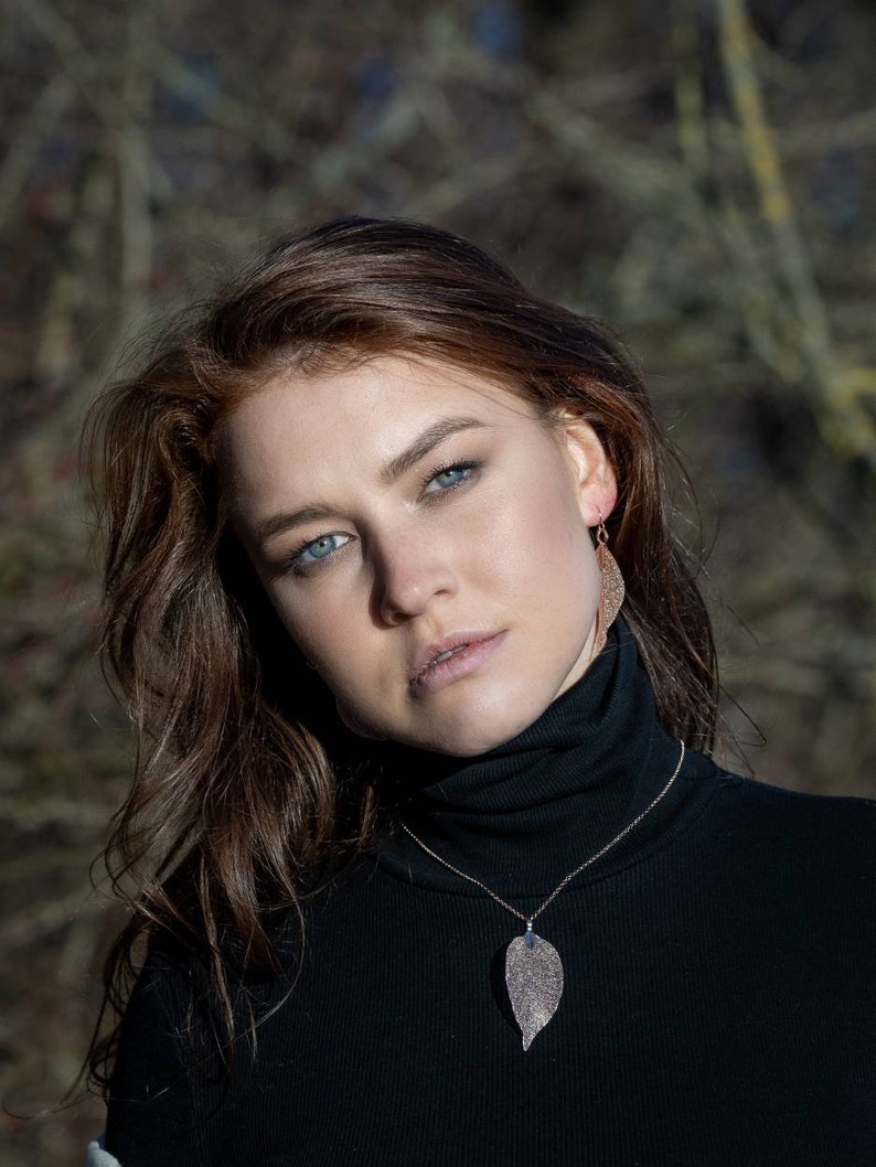 May include: A woman with long brown hair wearing a black turtleneck sweater with a gold necklace featuring a leaf pendant. The leaf pendant is made of a sparkly material.