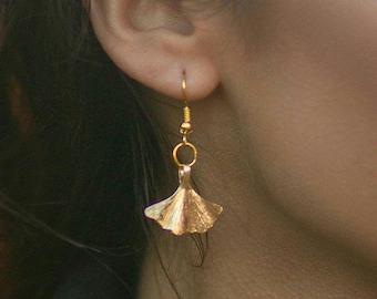 Puede incluir: Pendientes de hoja de ginkgo de oro con un diseño delicado. Los pendientes cuelgan de la oreja de una persona.