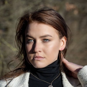 May include: A woman wearing a black turtleneck sweater with a gold chain necklace and a leaf pendant. She is also wearing a pair of leaf-shaped earrings. The woman has long brown hair and is wearing a white jacket.