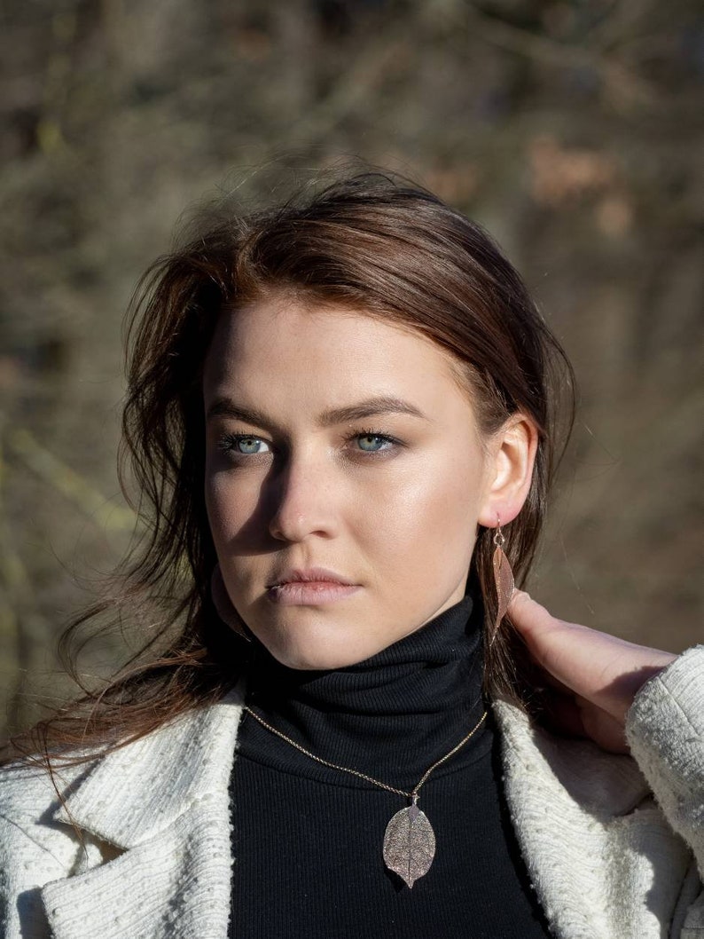 May include: A woman wearing a black turtleneck sweater with a gold chain necklace and a leaf pendant. She is also wearing a pair of leaf-shaped earrings. The woman has long brown hair and is wearing a white jacket.