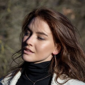 May include: A woman with long brown hair wearing a white jacket over a black turtleneck sweater. She is wearing a delicate gold necklace with a leaf pendant and matching leaf earrings.