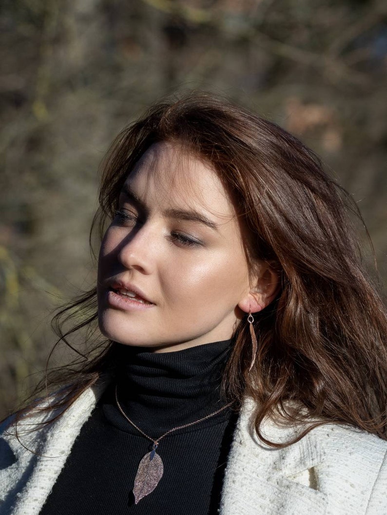 May include: A woman with long brown hair wearing a white jacket over a black turtleneck sweater. She is wearing a delicate gold necklace with a leaf pendant and matching leaf earrings.