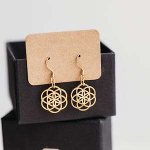 Peut inclure: Boucles d'oreilles pendantes dorées avec un motif géométrique de fleur de vie. Les boucles sont fixées sur un carton rectangulaire marron et présentées dans une boîte noire avec le texte SCHMUCK FEE GENNEY et un logo en forme de diamant.