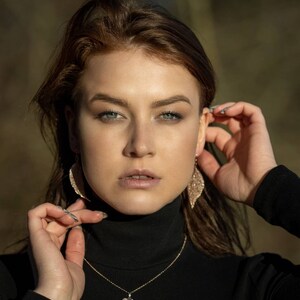 May include: A woman wearing a black turtleneck sweater with a gold chain necklace featuring a leaf pendant. She is wearing sparkly leaf-shaped earrings.