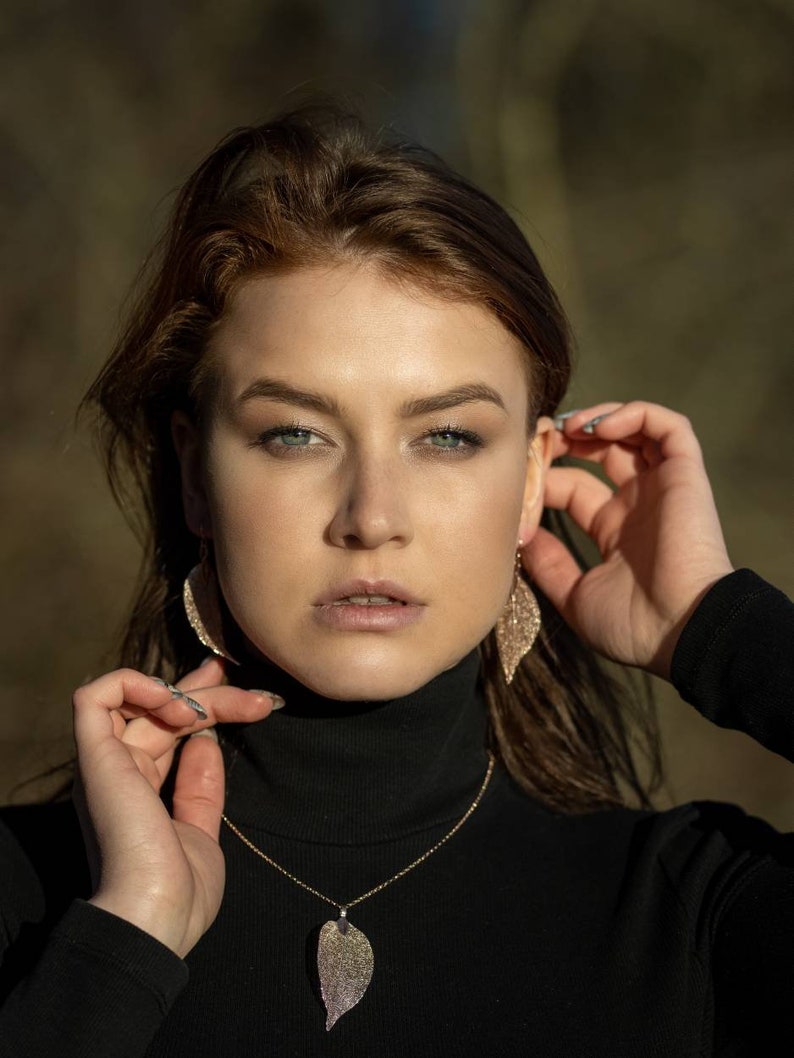 May include: A woman wearing a black turtleneck sweater with a gold chain necklace featuring a leaf pendant. She is wearing sparkly leaf-shaped earrings.