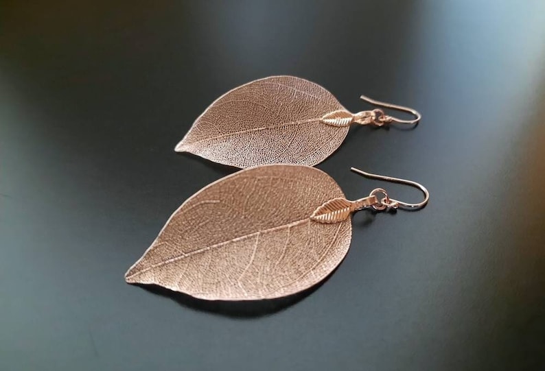 May include: A pair of rose gold leaf earrings. Each earring features a delicate, detailed leaf design with a small leaf charm hanging from the bottom.