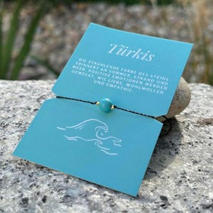Könnte beinhalten: Ein Armband mit einer türkisfarbenen Perle an einem schwarzen Band, präsentiert auf einer blauen Karte mit deutschem Text und einer Wellengrafik. Das Bild ist vor einem natürlichen Hintergrund im Freien aufgenommen.