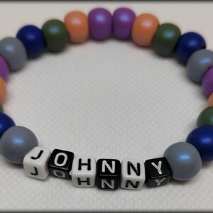 Könnte beinhalten: Ein farbenfrohes Perlenarmband mit dem Namen "JOHNNY", der in schwarzen Buchstaben auf weißen Perlen geschrieben ist.