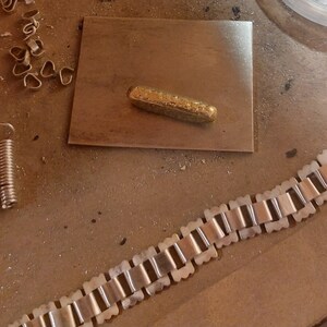 May include: A gold-colored metal bar on a gray surface, next to a gold-colored metal chain with square links.