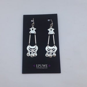 May include: A pair of silver dangle earrings with a geometric design and small silver balls hanging from the bottom. The earrings are displayed on a black card with the text "EPUWE PLATERIA MAPUCHE".