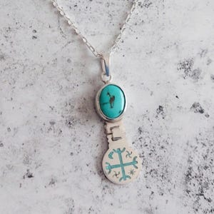 May include: A silver pendant necklace featuring a turquoise oval stone and a decorative cross design. The pendant hangs from a delicate silver chain. The turquoise stone is approximately 0.75 inches long.