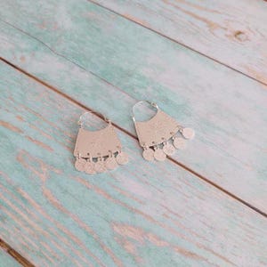 May include: A pair of silver hoop earrings with a decorative, scalloped design and dangling coin charms.