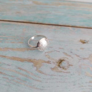May include: A silver ring with a round, embossed design featuring a flower motif. The ring is on a light blue wooden surface.