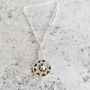 May include: A silver necklace with a pendant featuring a circular design with a central sphere and geometric patterns. The pendant is attached to a delicate silver chain.