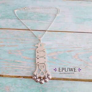 May include: A silver necklace with a detailed pendant featuring a geometric design and five small bells. The pendant is engraved with intricate patterns. The necklace is labeled "EPUWE PLATERIA MAPUCHE".