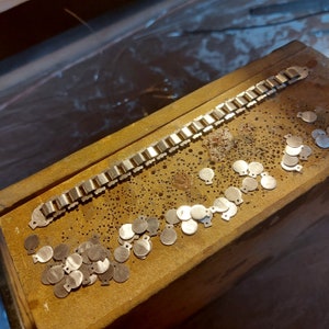 May include: A gold metal chain bracelet with a series of small, round, gold metal discs arranged in a pattern on a wooden surface.