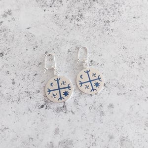 May include: A pair of silver earrings with a round pendant featuring a blue and white symbol. The symbol is a cross with four points, each with a crescent moon and a star.